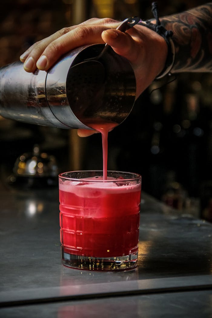 A bartender pours a vibrant red cocktail into a glass on a bar counter.